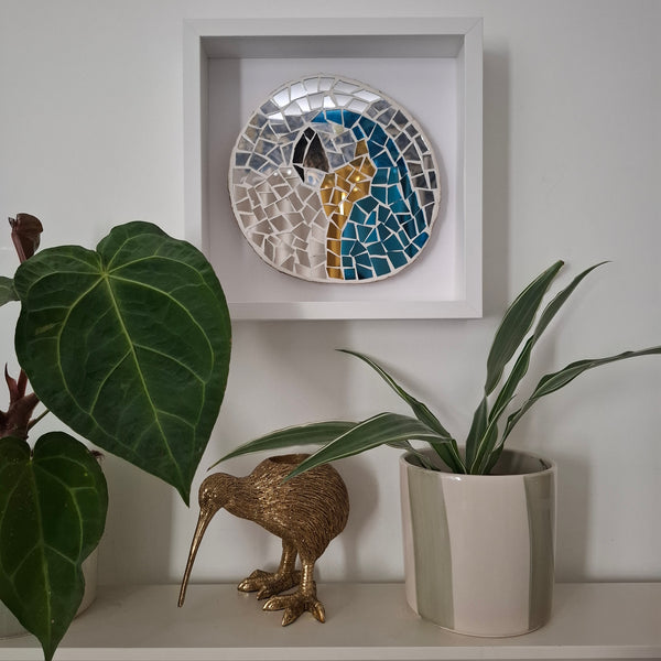 A round mosaic wall art piece featuring a stylized macaw in blue, gold, and white, mounted in a white square shadow box on a light gray wall. The display is styled on a shelf with green potted plants and a small gold kiwi bird figurine.