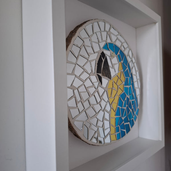 A circular mosaic wall art piece featuring a stylized bird design in blue, yellow, and white, mounted inside a white recessed frame.