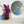 A round mosaic featuring a peacock design, styled on a white shelf next to two glass vases with purple and white flowers against a white wall.