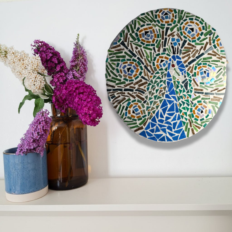 A round mosaic featuring a peacock design, styled on a white shelf next to two glass vases with purple and white flowers against a white wall.