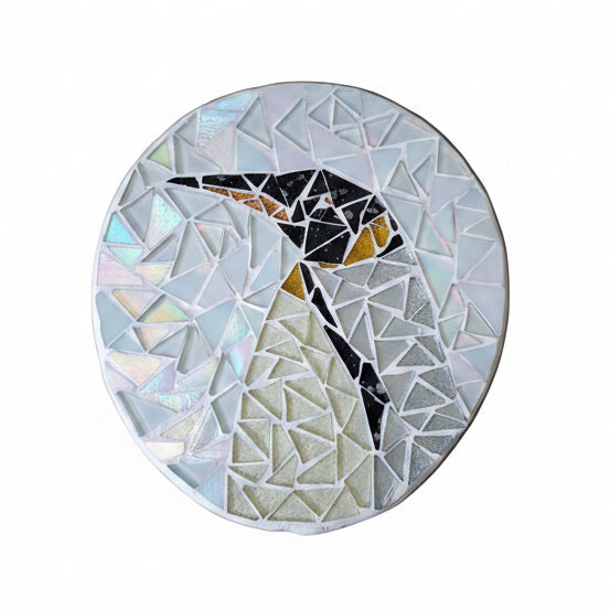 Circular mosaic wall art featuring a stylized penguin design. Crafted with glittery silver, black and white glass pieces on a frosted white background, set on rustic wood.