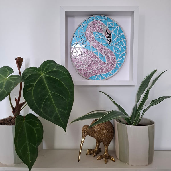 A framed circular mosaic wall art piece featuring a pink flamingo on a blue geometric background, displayed on a shelf above potted plants and a small kiwi bird figurine.