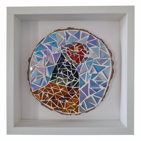 A square white shadow box frame containing a colorful mosaic bird artwork made from blue, purple, red, orange, green, and black glass pieces on a wood slice base.