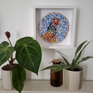 A square white shadow box frame displaying a colorful mosaic bird artwork, styled on a shelf with potted plants and a brown glass bottle against a white wall.