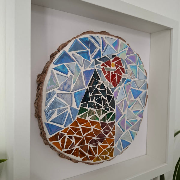 A circular mosaic wall art piece depicting a stylized bird, mounted on a rustic wood slice and displayed inside a white shadow box frame.