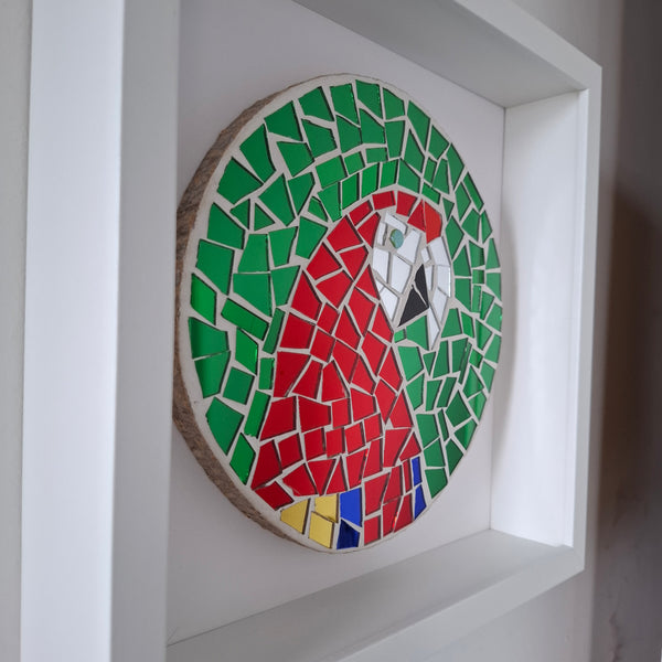 A circular mosaic wall art piece depicting a parrot, mounted inside a white frame. The parrot design features green, red, white, yellow, and blue mosaic tiles.