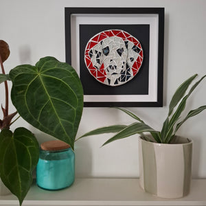 Framed circular mosaic wall art featuring a stylized dog face in red, white, black, and silver geometric shapes, displayed on a shelf above green houseplants.