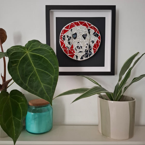 Framed circular mosaic wall art featuring a stylized dog face in red, white, black, and silver geometric shapes, displayed on a shelf above green houseplants.
