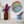 A round mosaic wall art piece with a rainbow geometric pattern hangs on a white wall above a shelf. On the shelf, an amber glass vase holds white and purple flowers, and a blue speckled ceramic pot holds purple flowers.