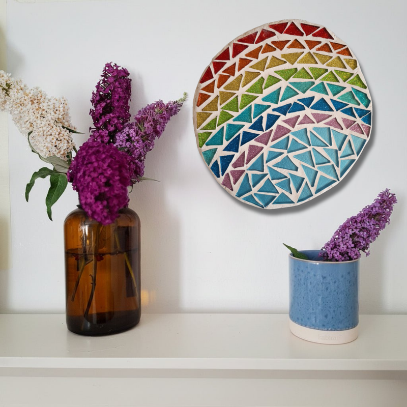 A round mosaic wall art piece with a rainbow geometric pattern hangs on a white wall above a shelf. On the shelf, an amber glass vase holds white and purple flowers, and a blue speckled ceramic pot holds purple flowers.