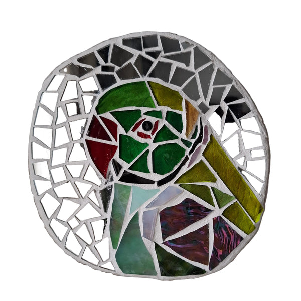 Circular mosaic wall art featuring a stylized parakeet design. Crafted with green stained glass pieces on a reflective silver mirror background, set on rustic wood.