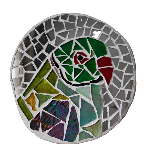 Circular mosaic wall art featuring a stylized parakeet design. Crafted with green stained glass pieces on a reflective silver mirror background, set on rustic wood.