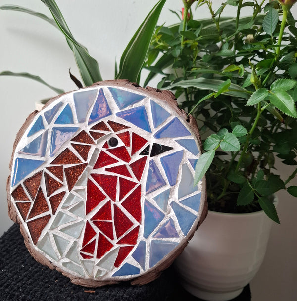 Circular mosaic wall art featuring a stylized robin design. Crafted with white, red, and brown glittery glass pieces on an iridescent blue background, set on rustic wood. Displayed on a table in a room with plants to show how beautiful the piece is in a home. 