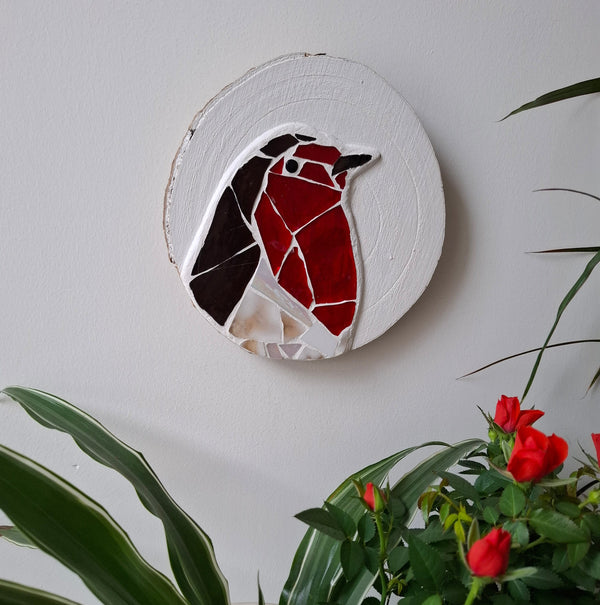 Circular mosaic wall art featuring a stylized robin design. Crafted with white, red, and brown stained glass pieces on a painted white background, set on rustic wood. Displayed on a wall in a room with plants to show how beautiful the piece is in a home. 