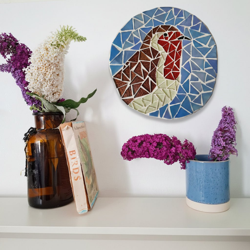 A round mosaic wall art piece featuring a robin displayed on a white wall above a shelf with flowers and books.