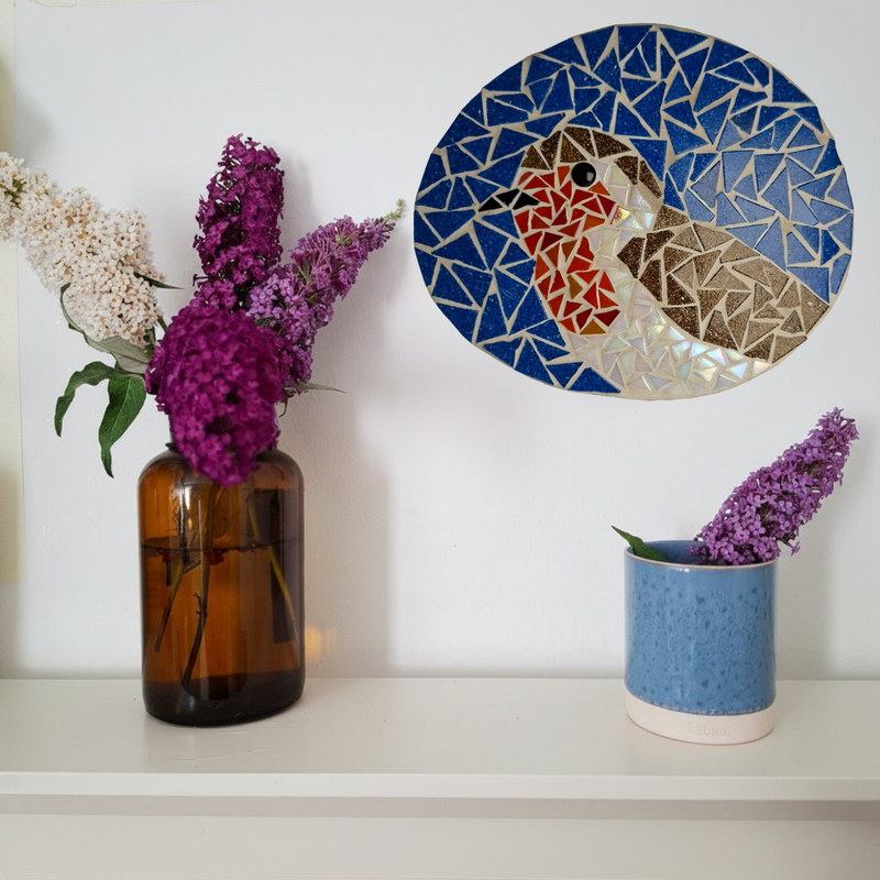 A styled interior scene with a  robin mosaic against a white wall. On the left is an amber glass vase holding purple and white flowers.