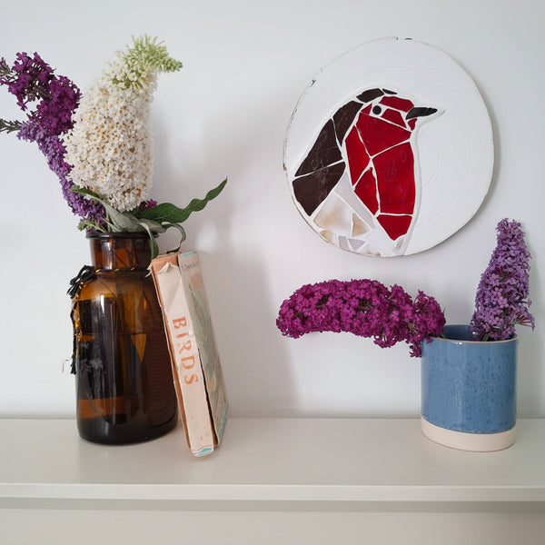 A styled interior scene with a white shelf against a white wall: an amber glass vase holding mixed dried flowers, a vintage book labeled BIRDS, a round stained-glass-style bird wall piece, and a small blue ceramic pot with purple flowers.