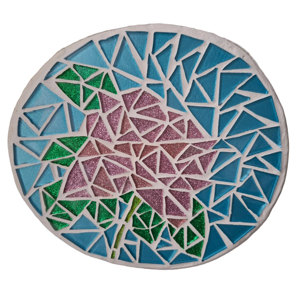 Circular mosaic wall art featuring a stylized rose design. Crafted with pink and green glittery glass pieces on a sparkling blue background, set on rustic wood.