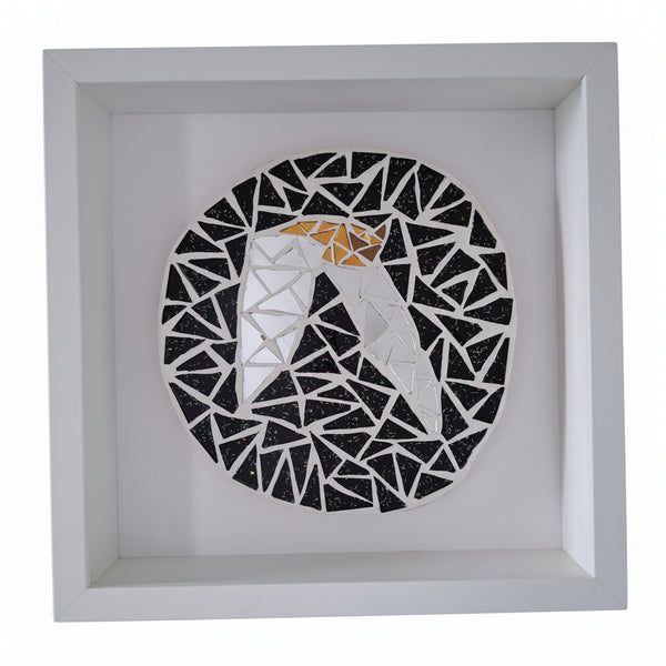 A square white shadow box frame containing a circular mosaic art piece of a stylized bat in black, silver and bronze.