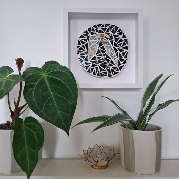 A circular black and silver mosaic wall art piece with a gold-toned bat at the center, displayed inside a white square shadow box on a white wall above a shelf with potted plants.
