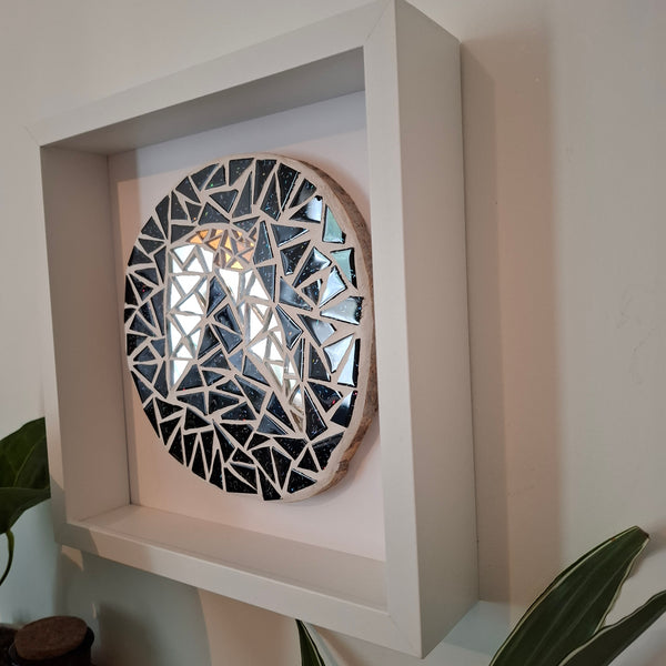 A circular mosaic wall mosaic with bronze and silver bat against a glittery black sky and a natural wood edge, displayed inside a white square shadow box mounted on a white wall.