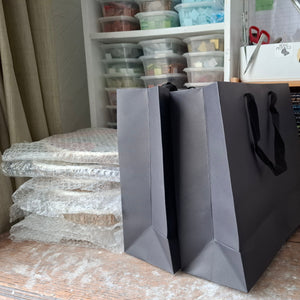 Two large black paper shopping bags with black handles standing on a wooden desk in a mosaic studio with shelves and supplies in the background.
