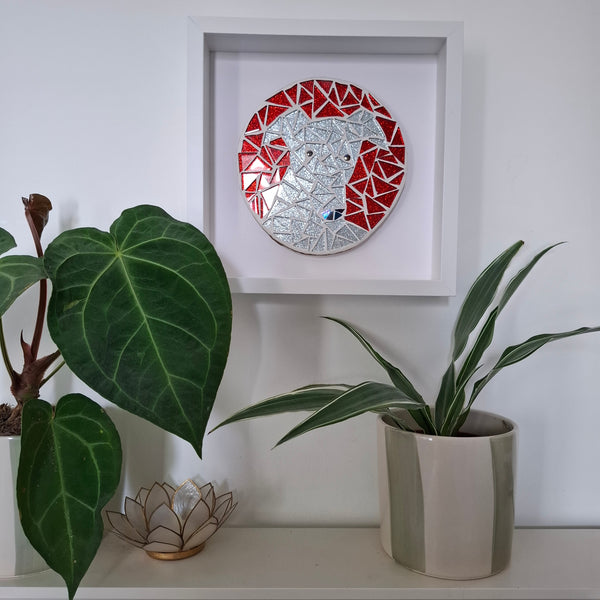A circular mosaic wall art piece featuring a dog's face in red and silver, displayed inside a white square frame on a white wall above a shelf with potted plants.