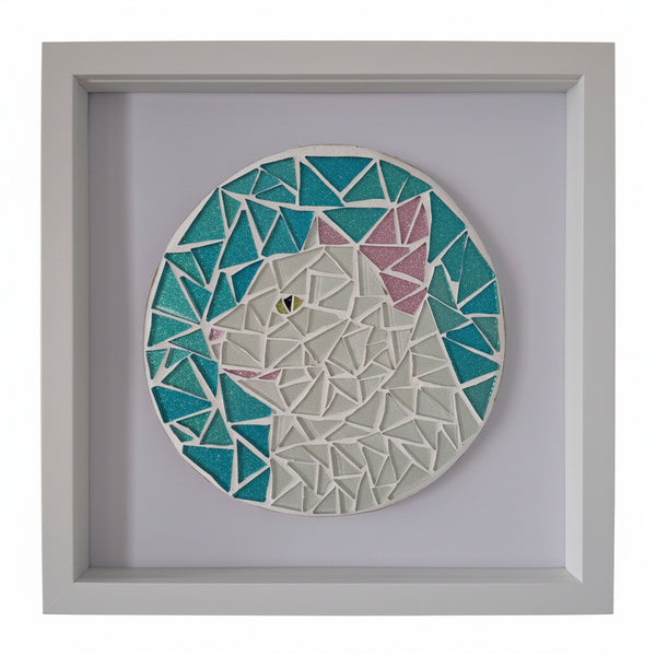 A square framed mosaic wall art piece depicting a stylized cat head in geometric triangular shapes. The artwork features teal blue, white, and pink sections, set within a light gray frame against a pale wall.