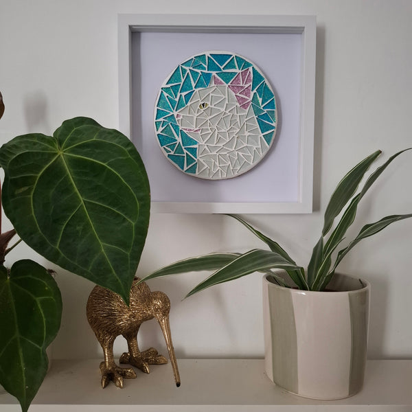 A framed mosaic cat wall art piece hangs on a white wall above a shelf with green plants and a gold kiwi figurine. The mosaic features a white cat with pink ears, surrounded by teal, blue, and white geometric tiles.