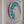 A circular mosaic wall art piece featuring a stylized cat face in shades of teal, blue, white, and soft pink, mounted on a white backing inside a white frame.