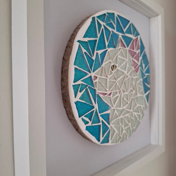 A circular mosaic wall art piece featuring a stylized cat face in shades of teal, blue, white, and soft pink, mounted on a white backing inside a white frame.
