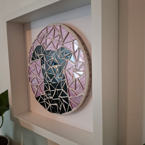 A circular mosaic wall art piece mounted inside a white frame. The artwork features a black greyhound on a pink background, with a natural wood edge.