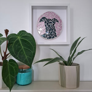A circular mosaic wall art piece of a black greyhound with pink background, displayed inside a white square frame on a white wall above a shelf with potted plants.