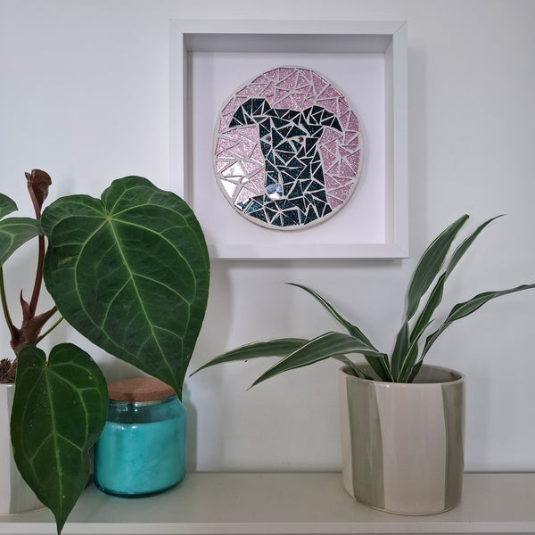 A circular mosaic wall art piece of a black greyhound with pink background, displayed inside a white square frame on a white wall above a shelf with potted plants.