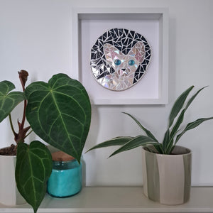 A circular mosaic wall art piece depicting a cat's face, mounted in a white square frame on a white wall above a shelf with potted plants.