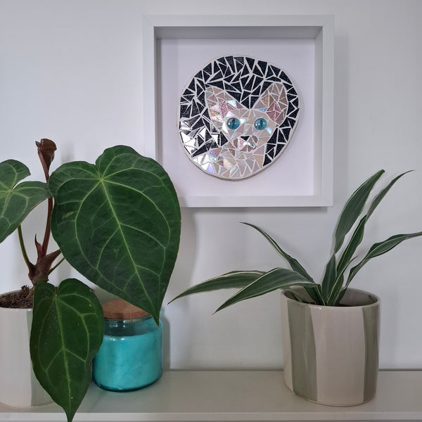 A circular mosaic wall art piece depicting a cat's face, mounted in a white square frame on a white wall above a shelf with potted plants.