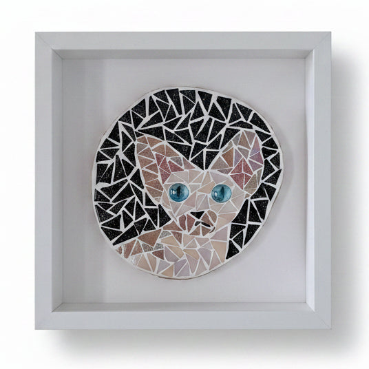 A circular mosaic wall art piece depicting a cat's face, mounted in a square white shadow box frame against a light gray wall.
