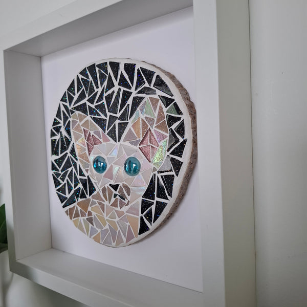 A circular mosaic wall art piece depicting a cat face, mounted inside a white square shadow box frame. The mosaic features black, white, beige, and pink tiles, with two prominent blue glass eyes.
