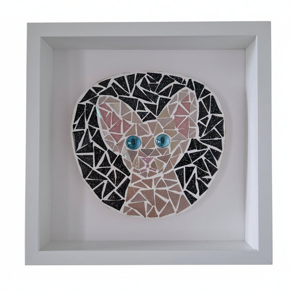 A square white shadow box frame containing a mosaic cat portrait. The cat design is made of black, beige, and pink triangular pieces with bright blue glass eyes, set against a white background.
