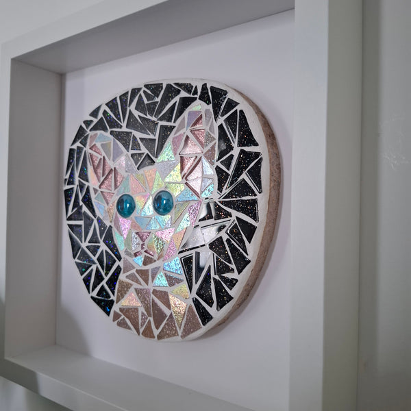 A circular mosaic wall art piece featuring a stylized cat face made from iridescent and black glass tiles, mounted inside a white frame.