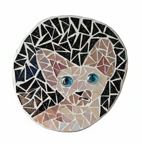 Circular mosaic wall art featuring a stylized cat design. Crafted with iridescent pink glass pieces on a sparkly black background, set on rustic wood.
