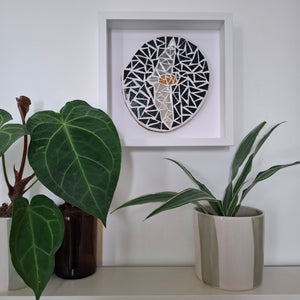 A circular black and silver mosaic wall art piece of a bat with outstretched wings in the center, displayed inside a white square shadow box frame on a white wall above a shelf with potted green plants.