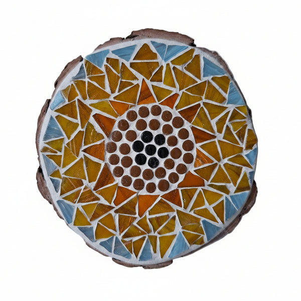 Circular mosaic wall art featuring a stylized sunflower design. Crafted with orange and yellow gold-dusted glass pieces on a pale blue background, set on rustic wood.
