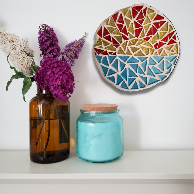 A decorative circular mosaic wall hanging with a geometric sunburst pattern in red, gold, and blue, displayed on a white wall above a shelf with an amber glass vase of purple flowers and a turquoise glass jar.