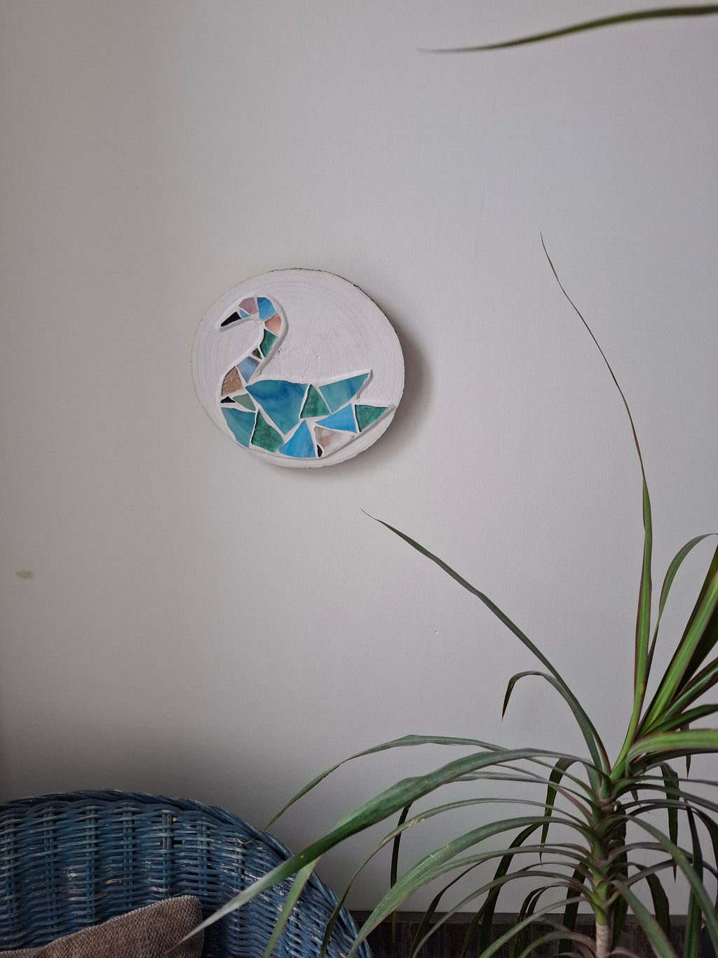 Circular mosaic wall art featuring a stylized swan design. Crafted with blue, green and aqua glass pieces and set on rustic wood. Displayed on a wall in a room with plants and a chair to show how beautiful the piece is in a home. 