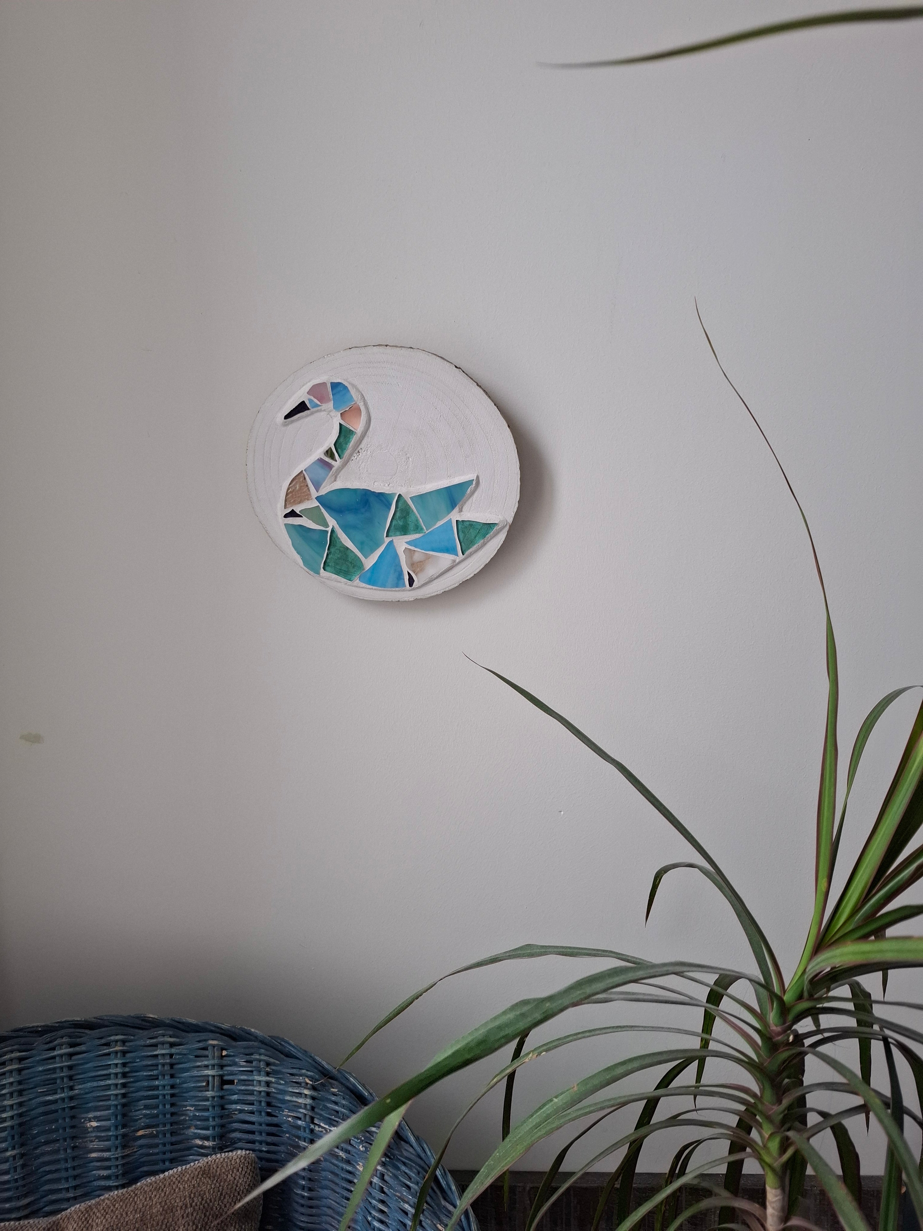 Circular mosaic wall art featuring a stylized swan design. Crafted with blue, green and aqua glass pieces and set on rustic wood. Displayed on a wall in a room with plants and a chair to show how beautiful the piece is in a home. 