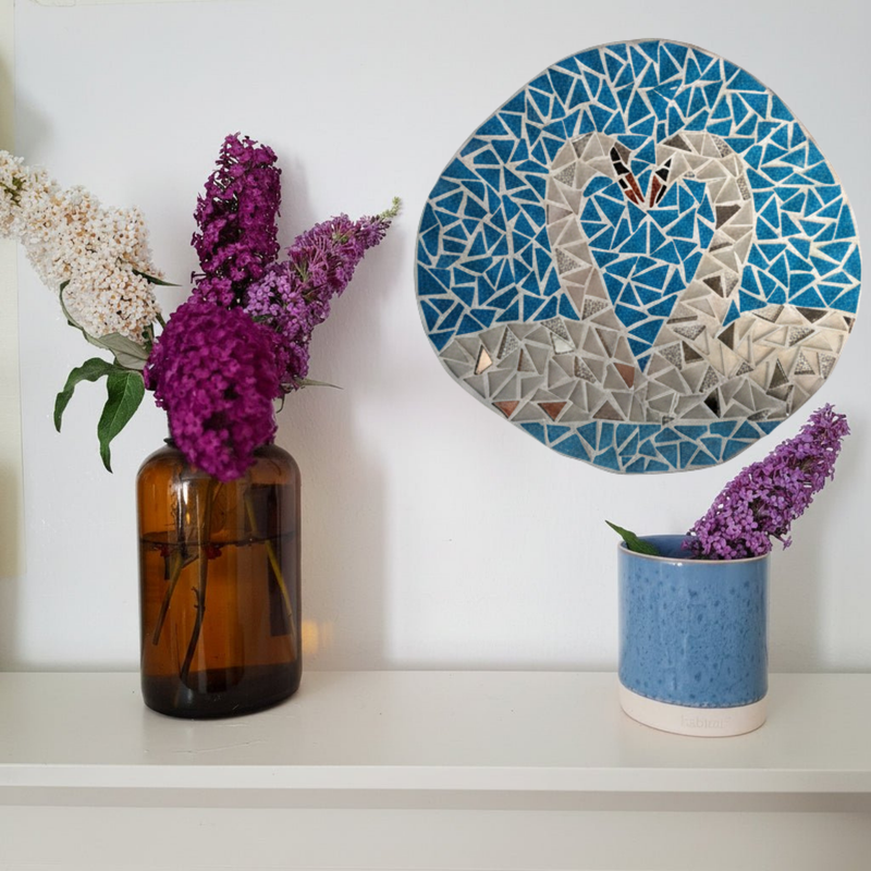 A decorative blue mosaic with a swan design hangs on a white wall above a shelf. The shelf holds an amber glass vase with white and purple flowers on the left and a small speckled blue pot with a purple flower on the right.