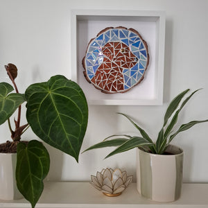 A square white shadow box frame displaying a colorful mosaic dog head artwork, styled on a shelf with potted plants and a small lotus-shaped decor piece against a white wall.