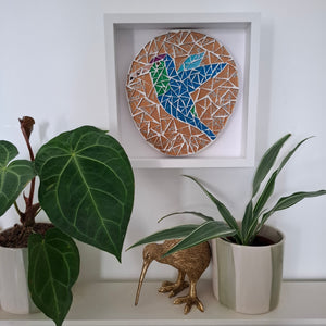 A circular mosaic wall art piece featuring a stylized hummingbird in blue, green and purple on a gold background. The mosaic is framed and hangs above a shelf with potted plants. 
