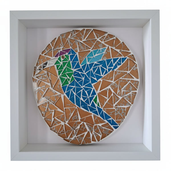 A circular mosaic bird wall art piece displayed inside a white square shadow box frame. The mosaic features a stylized bird constructed from blue, green, purple, and light blue tiles, set against a background of bronze and white geometric pieces.